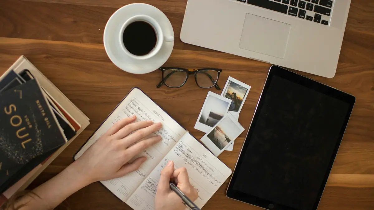 Someone writing in a notebook at a desk with coffee and a laptop