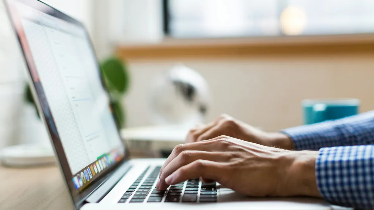Hands typing on a laptop on a wooden desk, with a plant and a mug in the background